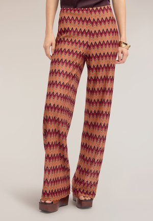 Vrouw draagt een broek met hoge taille en wijde pijpen met een oranje, rode en roze zigzagpatroon en bruine plateauzandalen.