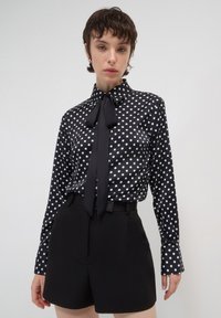 Schwarze Bluse mit weißen Pünktchen und einer Schleife am Kragen, kombiniert mit hochtaillierten, schwarzen Shorts, was einen stimmigen monochromen Look schafft.