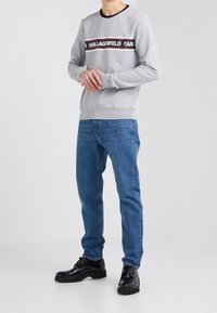 Sudadera gris con una banda de logo en negro y rojo cruzando el pecho, combinada con jeans azules y zapatos negros de cordones. Tela suave y ajuste cómodo.