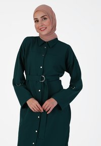Green button-up dress made of soft fabric, featuring a belted waist with a silver ring, long sleeves, and button accents down the front.