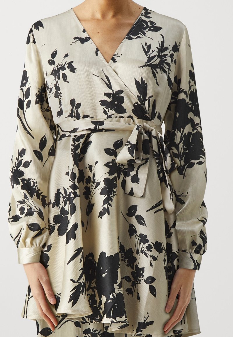 Robe portefeuille crème avec motif floral noir, manches longues bouffantes, col en V et ceinture nouée à la taille.