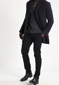 Mörkgrå ullkappa över en svart polo, kombinerad med svarta smala byxor och svarta ankellåga stövlar, med slät textur och skräddarsydd passform.