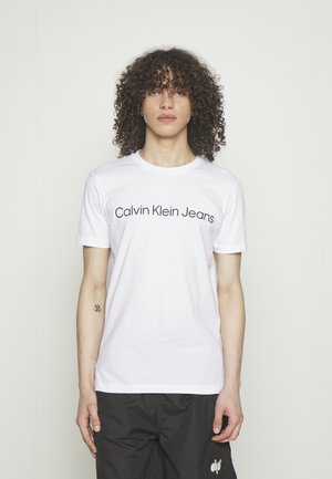 Osoba sa kovrčavom kosom koja nosi bijelu Calvin Klein Jeans majicu i crne hlače, stoji ispred jednobojne svijetle pozadine.