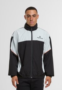 Juodas ir šviesiai mėlynas u�žtrauktukas striukė su aukštu apykakle, elastingomis rankovėmis ir laisvu siluetu. Logotipas yra išdėstytas ant krūtinės.