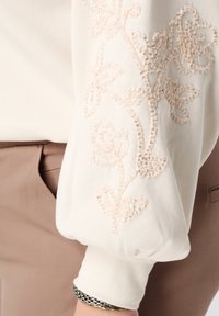 Witte pullover met opgepofte mouwen met beige bloemrijk borduursel en kralen. De stof oogt zacht, gecombineerd met beige broek.