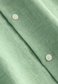 Tissu de chemise en lin vert clair avec une surface texturée, présentant deux boutons blancs. Le tissu tombe doucement avec des plis souples.