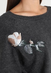 Ciemnoszary sweter dzianinowy z beżowymi, białymi i szarymi aplikacjami kwiatowymi z przodu, blisko dekoltu.