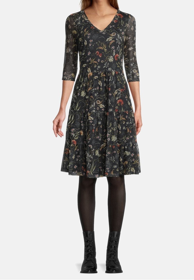 Mujer vistiendo un vestido negro con flores, de longitud a la rodilla y mangas tres cuartos, medias negras y botas de tobillo negras con cordones, de pie sobre un fondo blanco.