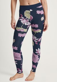 Donna che indossa dei leggings blu navy con un grande motivo floreale rosa e verde, in piedi scalza contro uno sfondo chiaro e semplice.