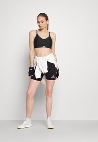 Schwarzer Sport-BH mit dünnen Trägern, schwarze Shorts mit weißen Akzenten und weiße Sportschuhe. Weißer Pullover um die Taille gebunden.