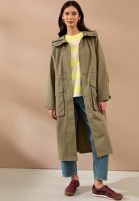 Langer olivgrüner Parka mit Reißverschluss, großen aufgesetzten Taschen und Kapuze, getragen über einem gelb gestreiften Hemd und blauen Jeans.
