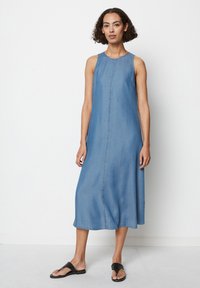 Lys blå denimkjole uten ermer med rett snitt, diskrete sømdetaljer og midilengde. Brukt med svarte flip-flops.