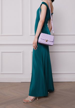 Vestido maxi ajustado color verde azulado con diseño de un solo hombro. Bolso acolchado color púrpura claro con herrajes dorados. Sandalias transparentes con tiras. Fondo de suelo de madera.