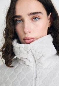 Jachetă din fleece gri deschis, cu guler înalt, model texturat în formă de diamond și închidere cu fermoar. Material moale cu un aspect pufos.