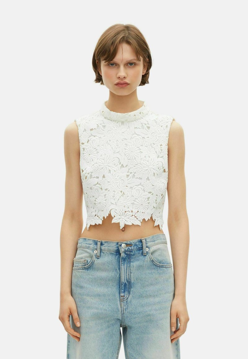 Jeune femme portant un haut court en dentelle florale blanche sans manches et un jean taille haute bleu clair sur fond uni.