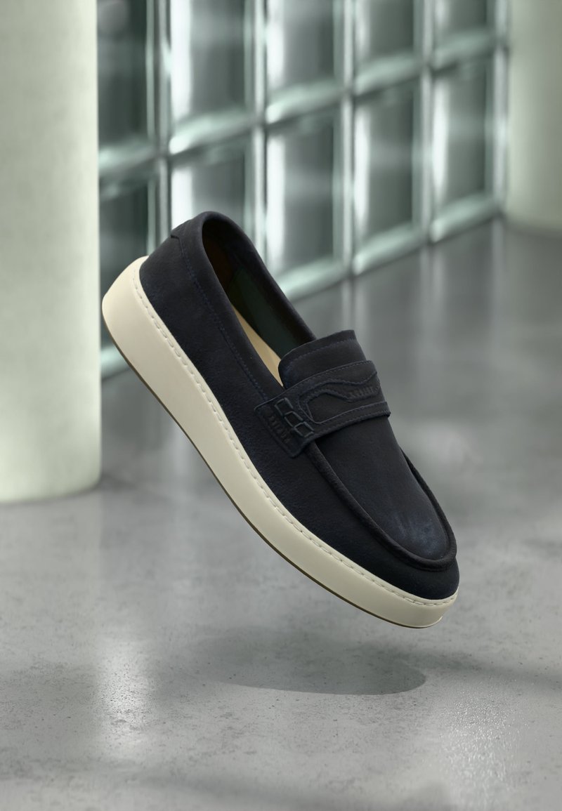 Mocassin en daim noir à semelle blanche flottant au-dessus d'un sol poli gris clair, avec un mur en blocs de verre et une colonne blanche en arrière-plan.