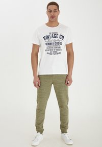 Bijela majica kratkih rukava s tamnoplavim printom na kojem piše "Vintage Co" u kombinaciji s maslinastozelenim trenirkama. Bijele tenisice upotpunjuju ovog outfita.