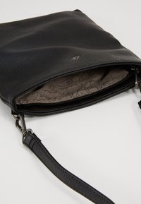 Černá kožená kabelka s texturovaným povrchem. Má zipové zapínání, stříbrný logo detail a vzorovanou látkovou podšívku uvnitř.