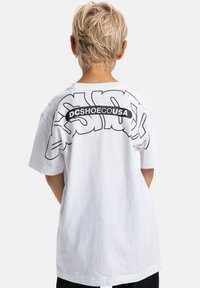 Biela bavlnená tričko s veľkým čiernym grafickým dizajnom s nápisom "DCSHOECOUSA" na chrbte. Krátke rukávy a uvoľnený strih.