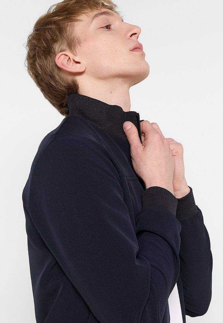 Chaqueta blanda en navy con cuello alzado, puños acanalados y cremallera en la parte delantera. Presenta una textura suave y un diseño minimalista.