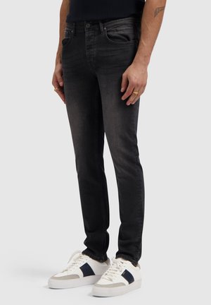 Man wearing slim-fit black jeans and white sneakers with black stripes, standing against a plain light background.