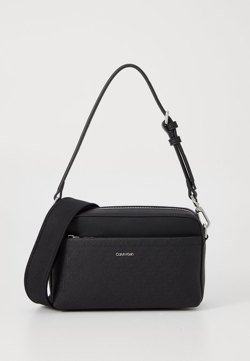 Sort crossbody taske med lynlås, struktureret materiale, justerbar rem og prægede logo-detaljer på forsiden.