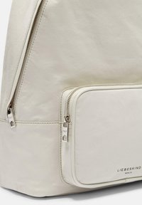 Zaino crema con un ampio scomparto principale e una piccola tasca frontale con zip. Caratterizzato da accessori in argento e una superficie liscia e strutturata.