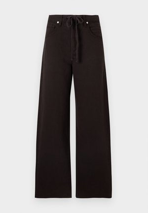 Moteris stovi, dėvi baltą ribotu megztinį su užtrauktuku, plačias juodas kelnes su raišteliais ir juodus aukštakulnius sandalus prieš paprastą foną.