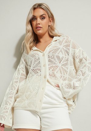 Crochet neuletakki vaaleassa valkoisessa, jossa on geometrinen kuvio, napitus ja väljä malli. Yhdistettynä valkoisiin shortseihin kerroksellista ilmettä varten.