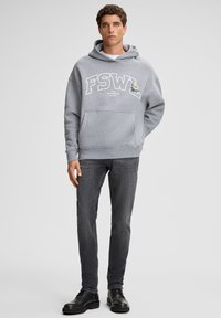 Giovane uomo che indossa un cappuccio grigio con il testo "FSWL", jeans grigio scuro e stivali neri, in piedi contro uno sfondo grigio chiaro semplice.