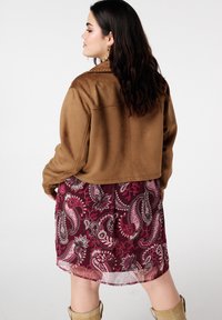 Veste courte en daim marron portée par-dessus une robe bordeaux à motif cachemire avec un tissu transparent. La tenue est complétée par des boucles d'oreilles en or et des bottes beige.