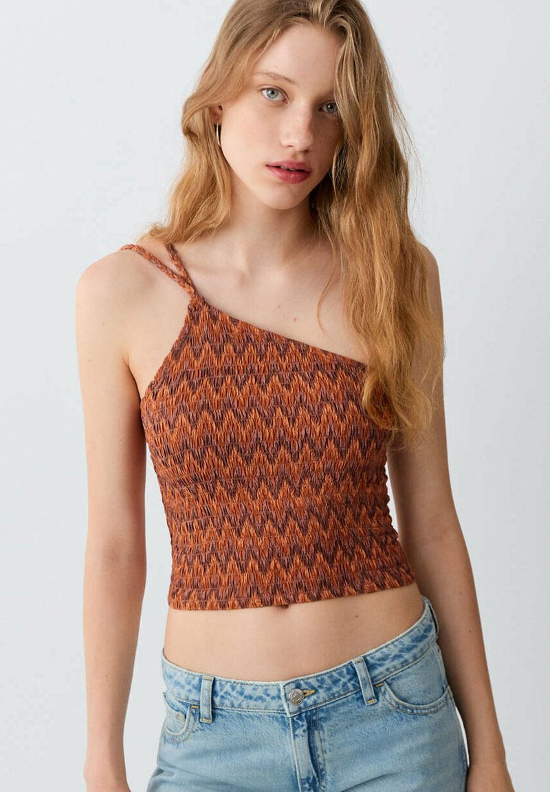 Mladá žena s dlouhými blond vlasy, která má oranžový vzorovaný crop top na jedno rameno a světle modré džíny na jednobarevném pozadí.