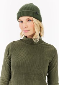 Olijfgroene geribbelde beanie en bijpassende coltrui. De beanie heeft een omgeslagen boord en een klein merketiket. Zachte, gestructureerde stof.
