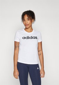 Bílé bavlněné tričko s krátkým rukávem, na přední straně s velkým černým logem "adidas", doplněné tmavě modrými legínami.