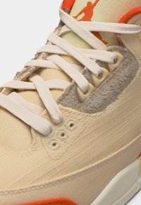 Κοντινό πλάνο κορδονιών μπεζ αθλητικού παπουτσιού δεμένα πάνω σε υφασμάτινη επιφάνεια με υφή, με πορτοκαλί λογότυπο Jumpman στην επένδυση της γλώσσας.