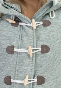 Světl zelená bunda s kapucí vyrobená z texturované látky, s dřevěnými zapínacími knoflíky a šedými semišovými detaily na místě zapínání.