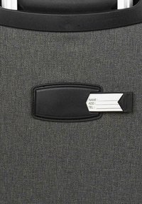 Maleta de tela gris con un soporte de etiqueta de identificación de cuero negro. La etiqueta está parcialmente extraída, presentando campos blancos para nombre, dirección y teléfono.