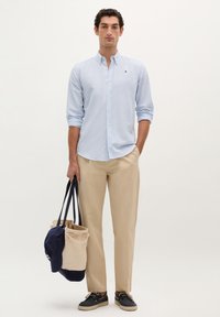 Lyseblå stribet skjorte med knapper, beige bukser og marineblåt fodtøj. Holder en lærredstotebag med en marineblå kasket ovenpå. Simpel, skræddersyet design.