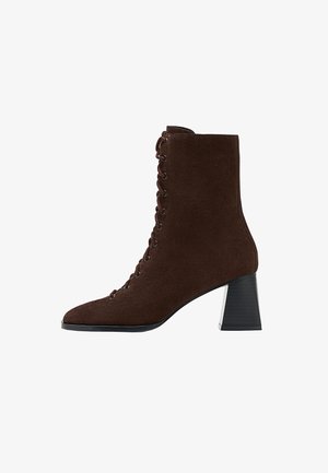 Brązowy zamszowy kowbojek sięgający kostki z ostrym noskiem, sznurowanym designem i grubym słupkowym obcasem. Charakteryzuje się teksturowanym materiałem oraz metalowymi oczkami.