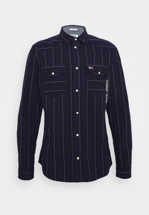 Navyblauw shirt met lange mouwen en witte verticale pinstripes, een knoopsluiting aan de voorkant, twee borstzakken met knopen en de tekst "Tommy Jeans" aan de rechterkant.