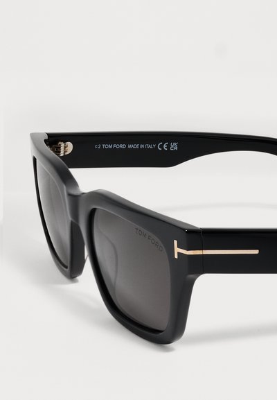 Lunettes de soleil carrées noires Tom Ford avec des verres foncés et un logo doré en forme de T sur la branche, marqué Made in Italy à l’intérieur de la branche.