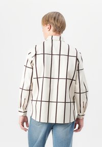 Camisa de algodão com base creme e padrões de quadrados castanhos audaciosos, colarinho, mangas longas e punhos com botões. Vista de costas.