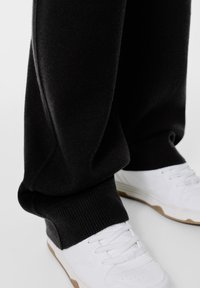 Fekete melegítőnadrág sima tapintású, gumis szegéllyel, fehér sportcipőkkel párosítva, amelyek texturált talppal és fűzővel rendelkeznek.