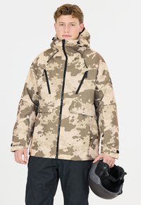 Vaaleanruskea camo-talvitakki, jossa on vetoketjullinen etuosa, mustat yksityiskohdat ja kaksi rintataskua. Mallilla on musta kypärä käsissään ja hänellä on päällään mustat housut.