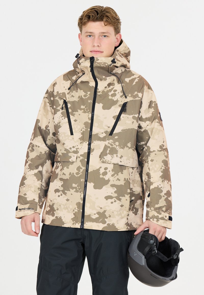 Vaaleanruskea camo-talvitakki, jossa on vetoketjullinen etuosa, mustat yksityiskohdat ja kaksi rintataskua. Mallilla on musta kypärä käsissään ja hänellä on päällään mustat housut.