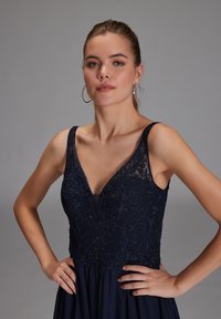 Abito da sera blu navy con scollatura a V, con corpetto in pizzo dettagliato con perline floreali e gonna fluente. Texture liscia, design senza maniche.