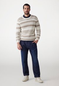 Leichter Pullover aus Creme mit horizontalen beigen Streifen, kombiniert mit dunkelblauen Hosen und cremefarbenen Sneakers. Der Pullover hat eine gerippte Textur.
