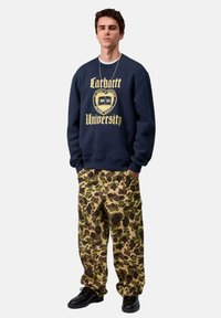 Sudadera de color azul marino con estampado amarillo de "Carhartt University"; pantalones de camuflaje en tono beige con parches oscuros; zapatos negros; el modelo se encuentra de pie contra un fondo blanco.