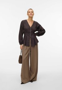 Dunkelbraune Bluse mit langen Ärmeln und V-Ausschnitt, die mit einer Reihe von Knöpfen auf der Vorderseite versehen ist. Kombiniert mit weiten beigen Hosen und einer kleinen Tasche.
