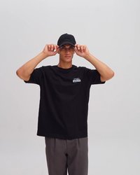 Camiseta negra con el logotipo "STUDIOS", de manga corta y corte relajado; combinada con una gorra de béisbol negra. El modelo posa con las manos sobre la gorra.